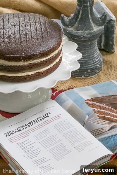 A stunning triple-layer chocolate cake with a creamy vanilla buttercream filling, partially unfrosted, sitting elegantly on a cake stand. An open cookbook lies nearby, inviting bakers to dive into the recipe. The image showcases the beautiful layers and the smooth texture of the buttercream.
