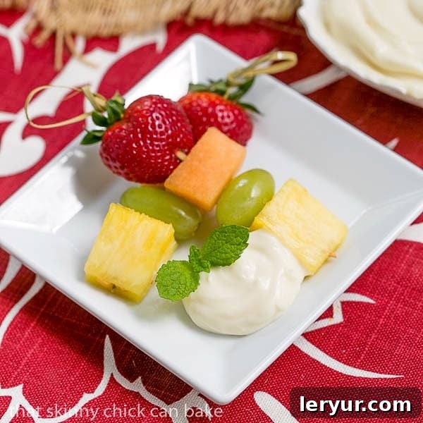 Rainbow Skewers with White Chocolate Mascarpone Dip - A dreamy dip to accompany vibrant fresh fruit kebabs A single fruit kebab resting on a small square plate next to a dollop of white chocolate mascarpone dip, garnished with a sprig of fresh mint.