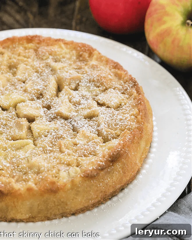 A whole, uncut French Apple Cake beautifully presented on a white cake plate, showcasing its golden-brown crust and rustic charm.