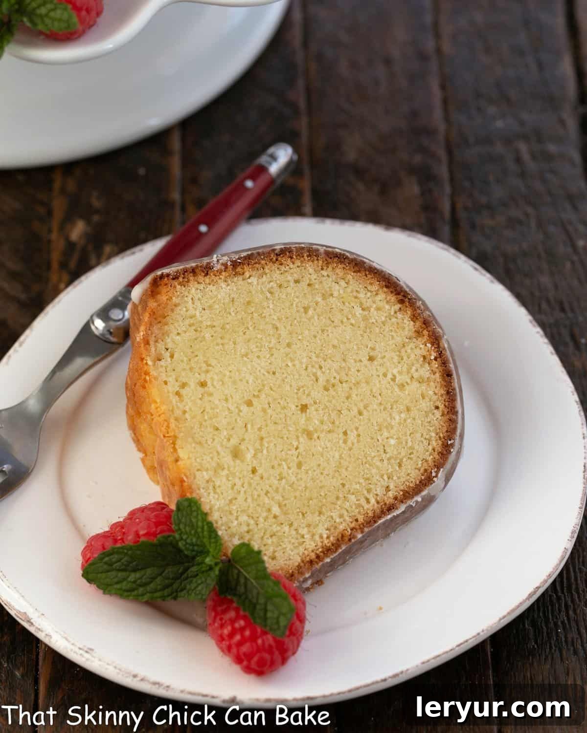 A perfectly baked slice of sour cream pound cake, elegantly presented on a white dessert plate with a red-handled fork and garnished with vibrant fresh raspberries, inviting indulgence.