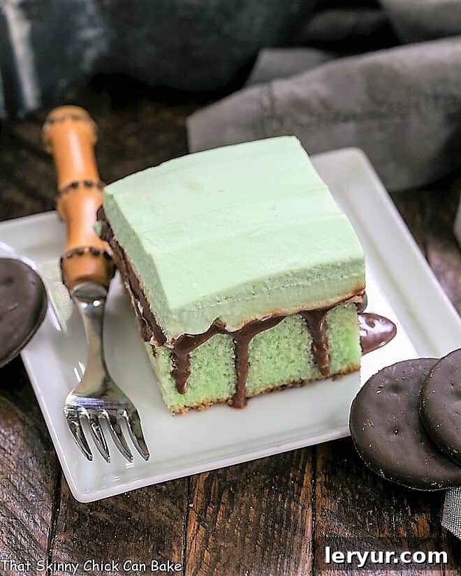 Creme de Menthe Cake - Mint infused cake topped with fudge and minty whipped cream A perfectly portioned slice of Creme de Menthe cake, showcasing its vibrant green layers, rich fudge, and creamy mint topping, artfully placed on a square white plate with a bamboo fork ready for tasting.