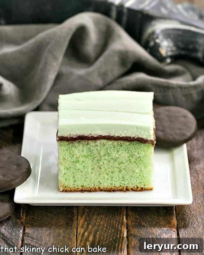 Creme de Menthe Cake - Mint infused cake topped with fudge and minty whipped cream An enticing close-up of a slice of grasshopper cake on a square white plate, showing the distinct layers of green mint cake, rich fudge, and fluffy mint cream.