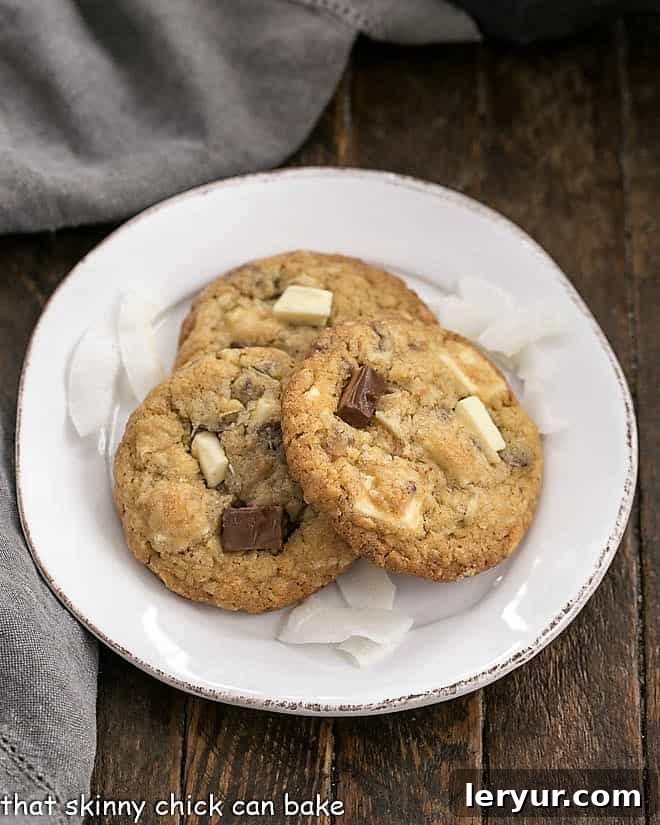 Chewy Oatmeal Toffee Cookies with a coconut bonus - fabulous flavors and textures in every bite make for truly irresistible cookies! Three golden-brown Chewy Oatmeal Toffee Cookies, adorned with visible toffee bits and white chocolate chips, resting on a pristine white dessert plate, promising an irresistible bite.