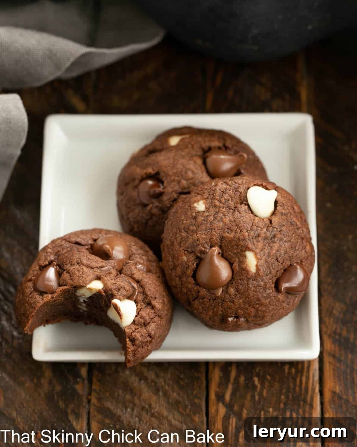 3 chocolate chocolate chip cookies on a square white plate, one with a bite removed.