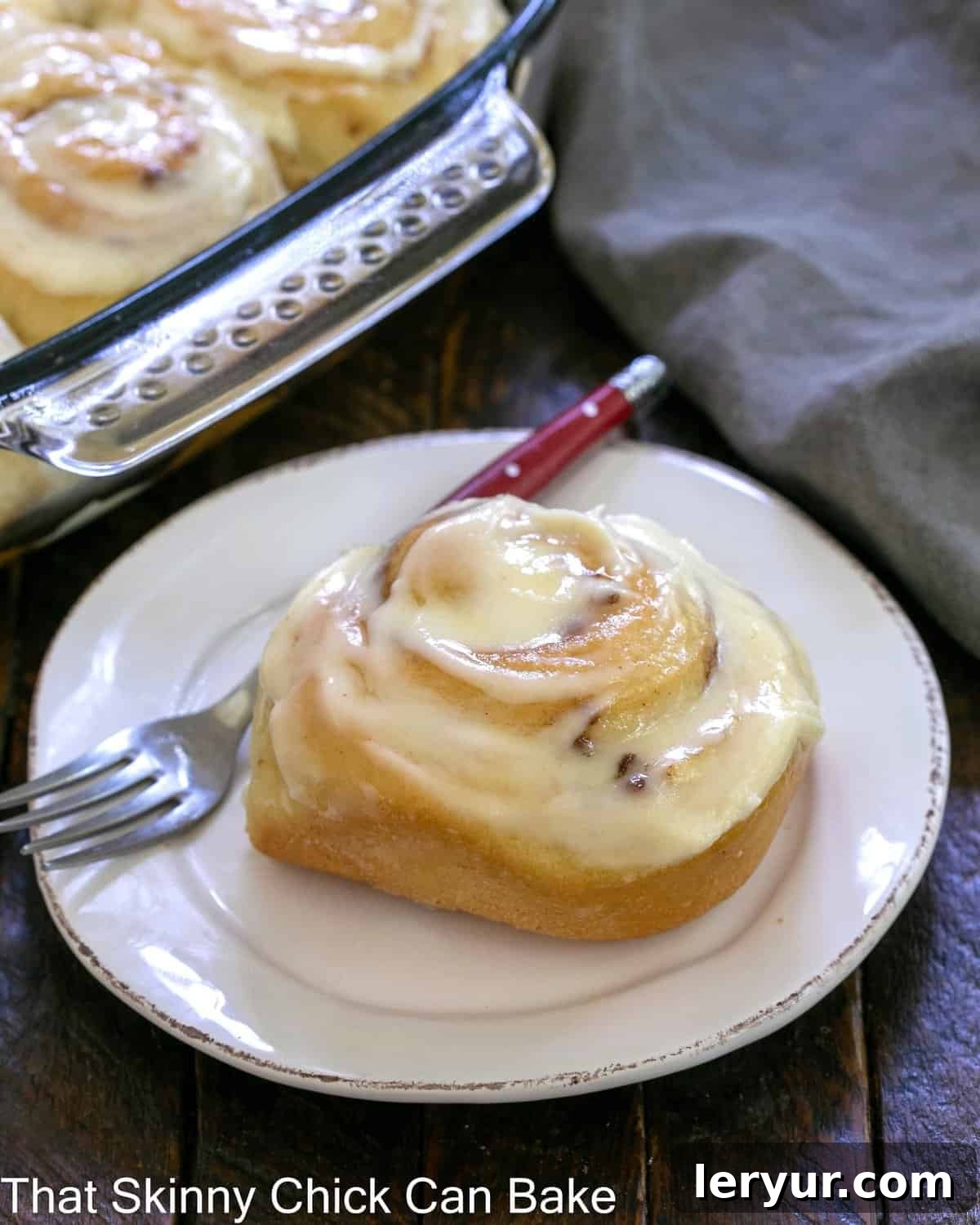 Grandma's Classic Cinnamon Buns 2 One perfectly frosted cinnamon roll on a white plate, with a pan of freshly baked rolls blurred in the background, highlighting its golden-brown exterior and generous vanilla icing.