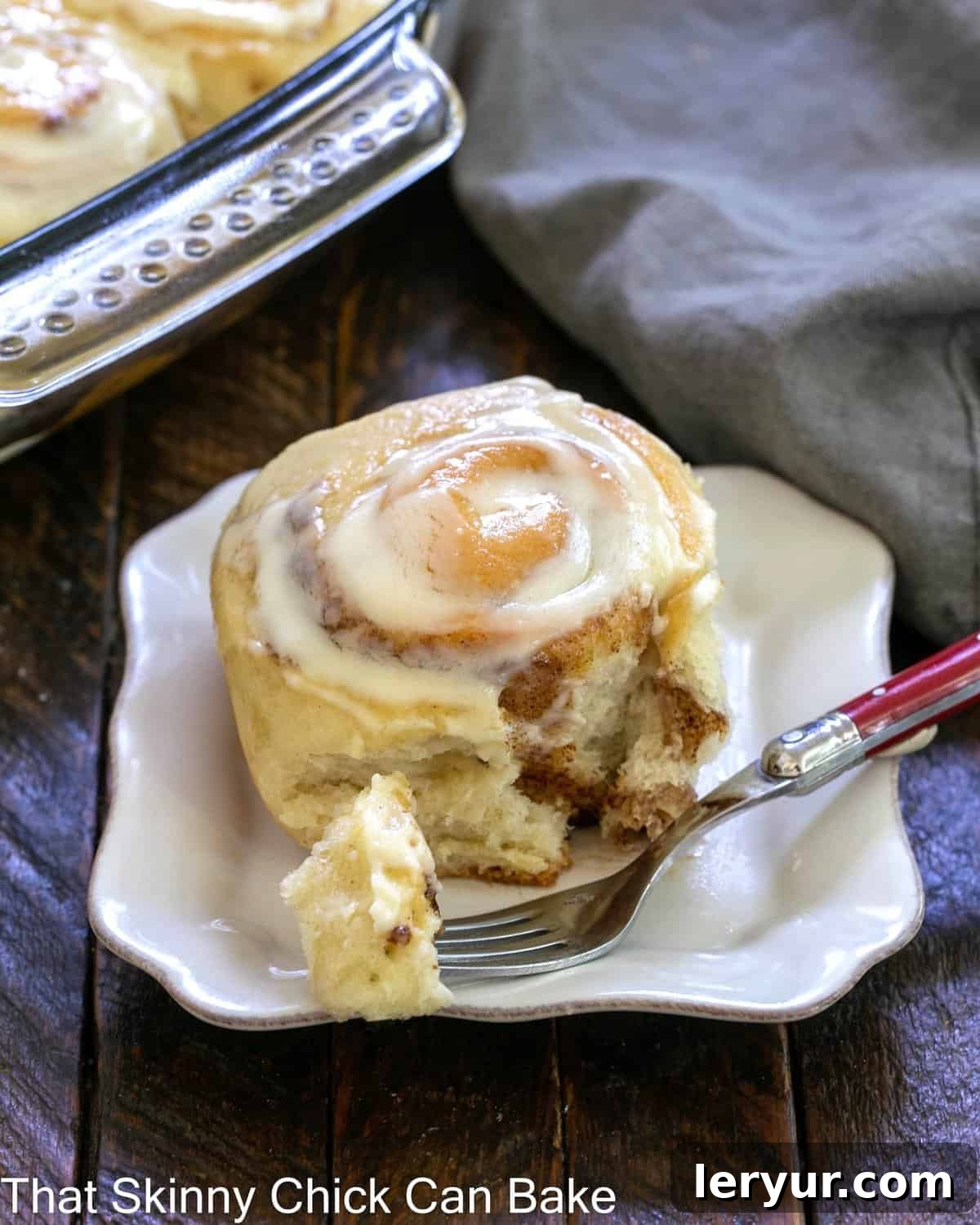 Old Fashioned Cinnamon Rolls - Tender with cinnamon swirls and vanilla icing A freshly baked cinnamon roll on a square white plate with a bite taken out, resting on a red-handled fork, revealing its perfectly swirled layers and creamy frosting.