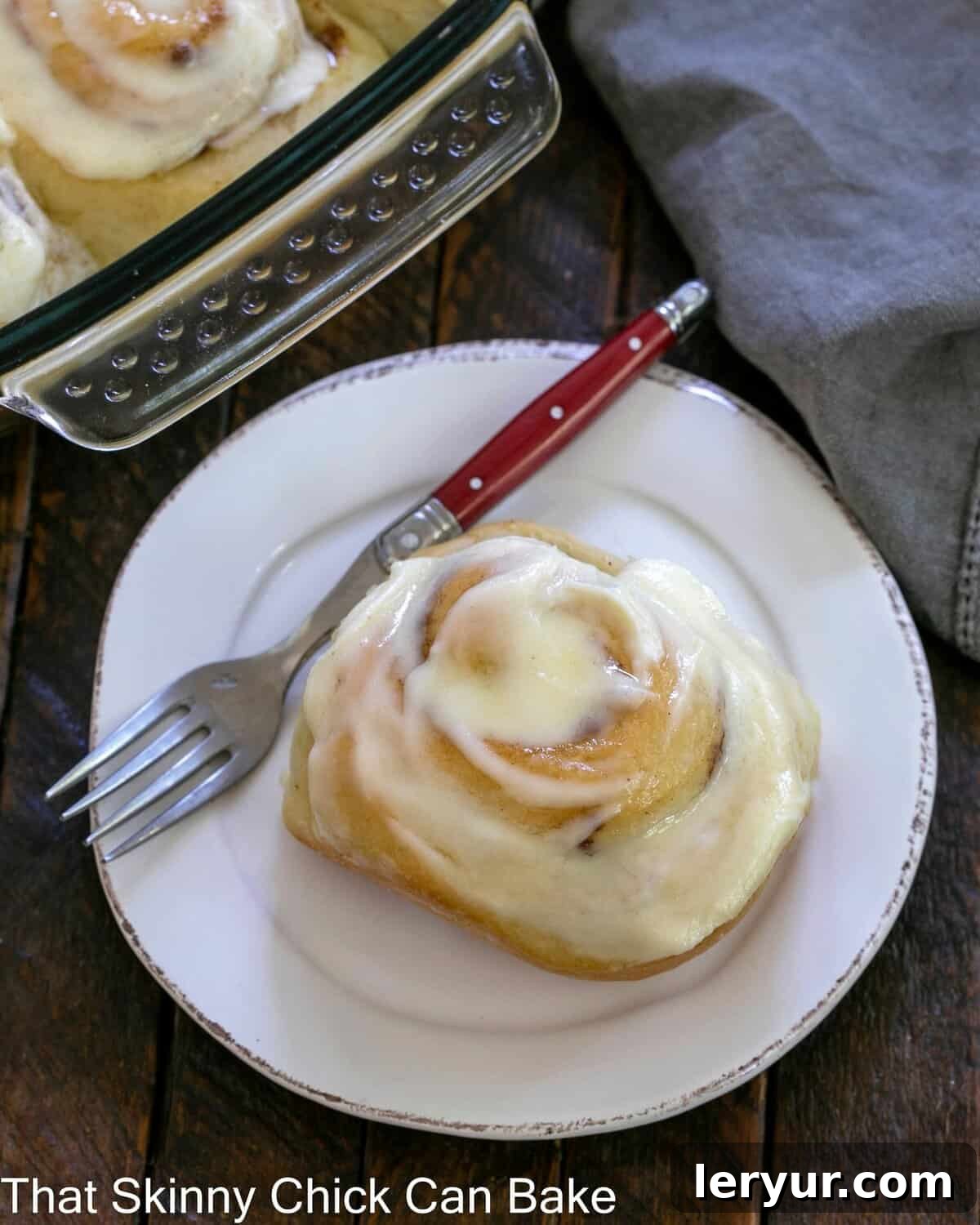 Old Fashioned Cinnamon Rolls - Tender with cinnamon swirls and vanilla icing An overhead shot of a single, perfectly frosted cinnamon roll on a round white plate, accompanied by a stylish red-handled fork, ready to be enjoyed.