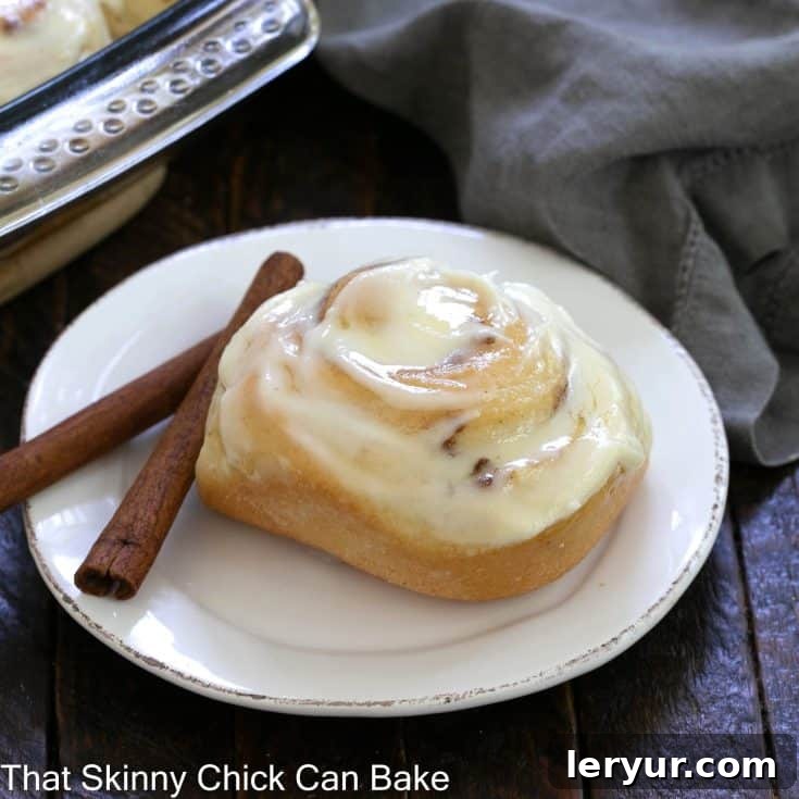 a frosted cinnamon roll on a round white plate with 2 cinnamon sticks