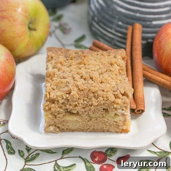 Apple Coffee Cake - A perfect blend of chopped apples, warm spices, and a delightful brown sugar cinnamon streusel. A delectable slice of Apple Coffee Cake on a white plate, beautifully arranged with fresh apples and cinnamon sticks in the background, inviting you to taste autumn's delight.