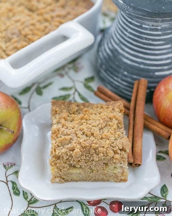 Apple Coffee Cake - A delightful composition of tender cake, sweet apples, warm spices, and a crunchy brown sugar streusel. A close-up of a generous slice of Apple Coffee Cake, showcasing its moist interior, apple chunks, and golden streusel topping, served on a modern square white plate with decorative cinnamon sticks.
