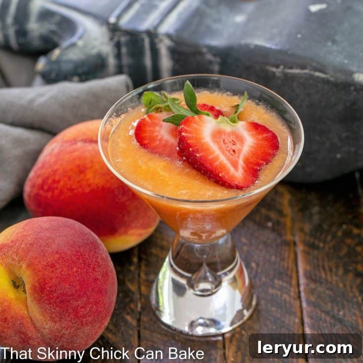 A peach bellini cocktail topped with sliced strawberries, next to two fresh peaches.