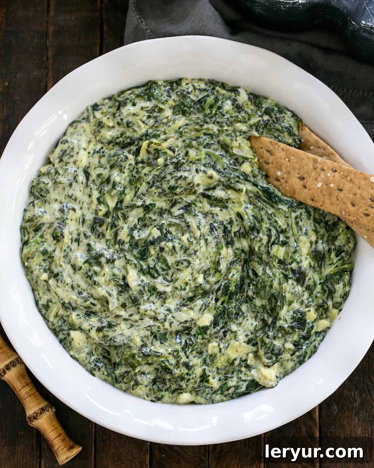 The ultimate warm Cheesy Spinach Dip recipe, perfect for parties and game day. Loaded with spinach, artichoke hearts, and cheese. Find the best spinach dip recipe here! Overhead view of a white casserole dish filled with warm and bubbly Cheesy Spinach Dip.