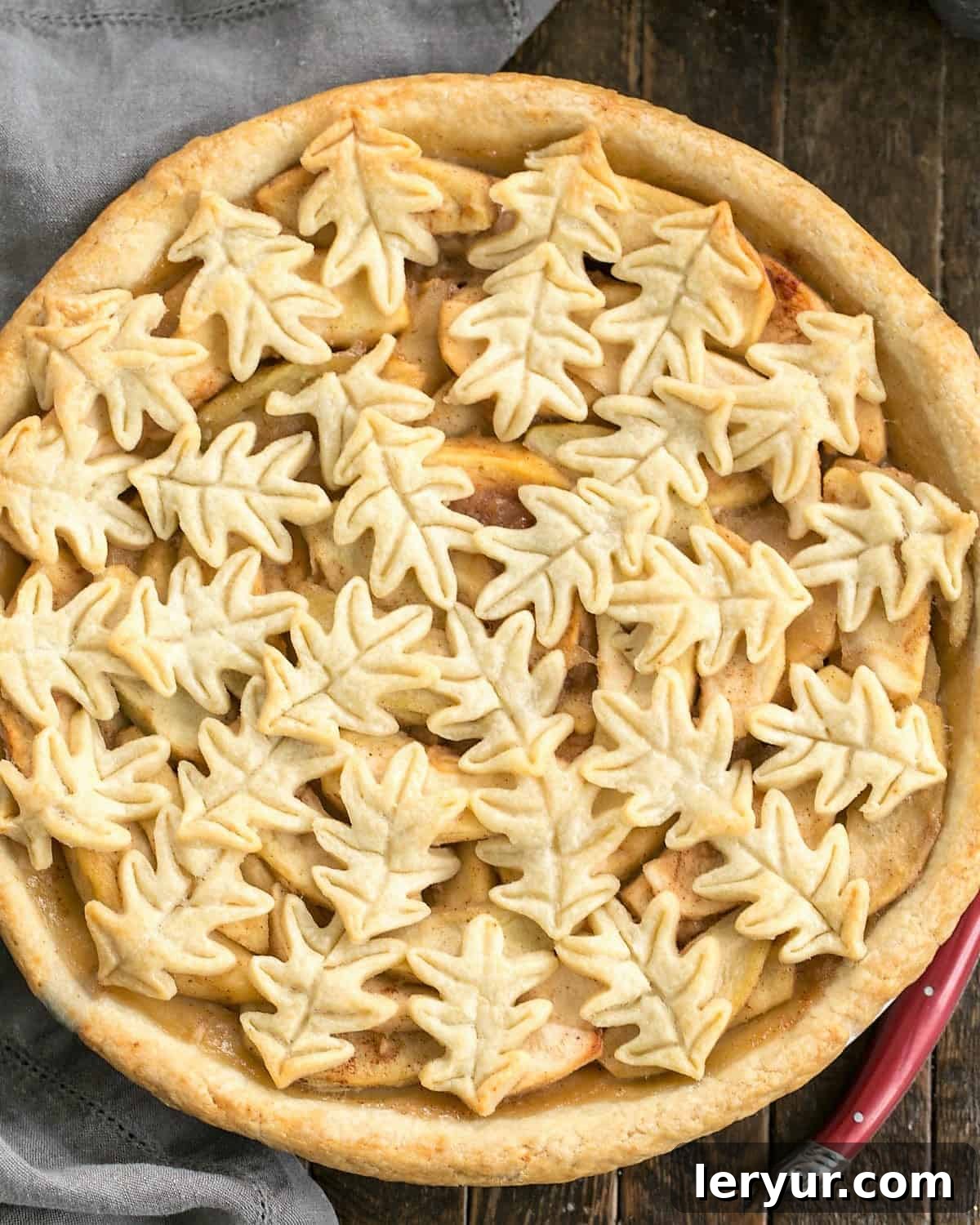 Overhead view of a freshly baked apple pie with an intricate leaf topping, next to a red-handled knife.