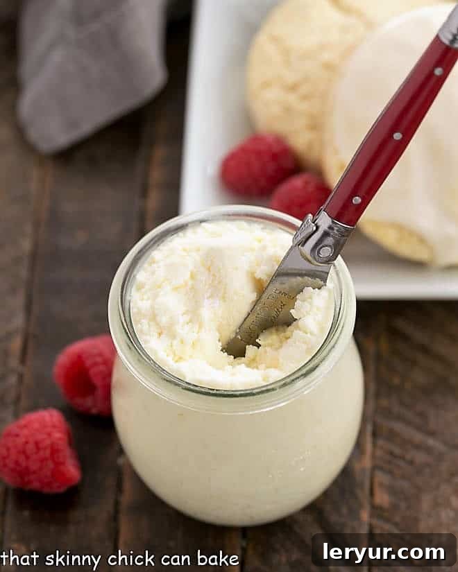 A jar of freshly made Instant Pot Clotted Cream, surrounded by fresh berries, fluffy scones, and a red-handled knife.