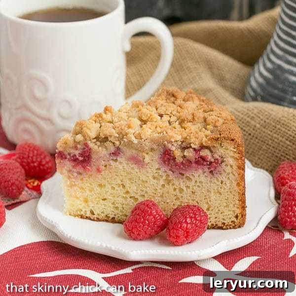 A slice of raspberry coffee cake on a white plate with fresh raspberries and a fork, showing the moist crumb and streusel topping