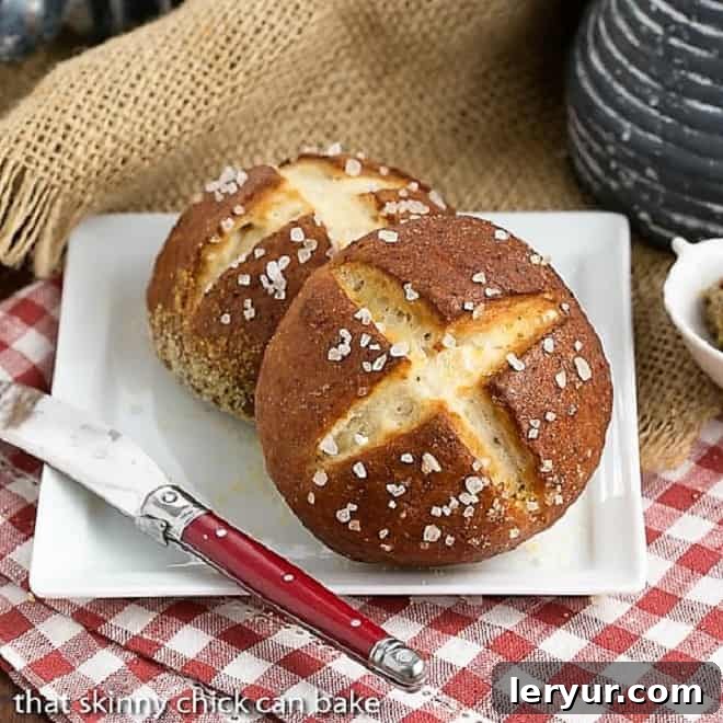 Two golden brown Homemade Pretzel Rolls, perfectly scored with an X, resting on a crisp white square plate beside a red-handled knife. The image captures the inviting texture and color of freshly baked bread.