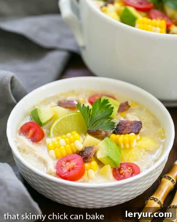 A charming white bowl filled with creamy chicken corn chowder, with a large cooking pot blurred in the background.
