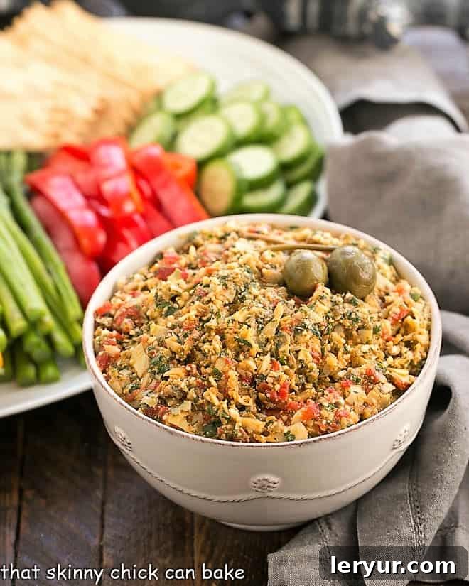 Red Pepper Artichoke Tapenade - a marvelous Mediterranean dip that's fabulous served with crackers or fresh vegetables A beautiful white bowl brimming with vibrant Roasted Red Pepper Artichoke Tapenade, surrounded by a colorful platter of fresh vegetables like cucumber slices, carrot sticks, bell pepper strips, and cherry tomatoes, ready for dipping.