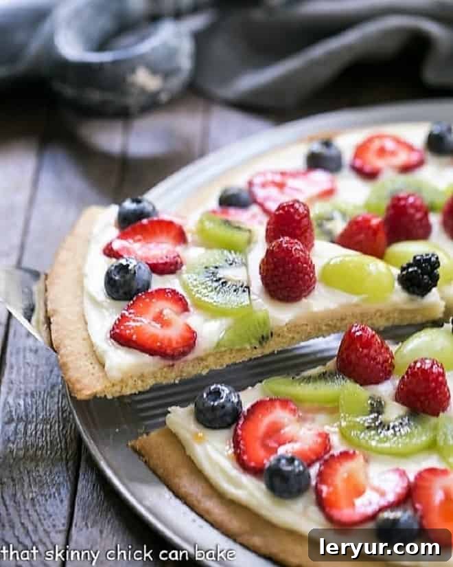 A vibrant slice of Fresh Fruit Pizza being carefully lifted from a larger pizza on a sleek metal pan, showcasing its colorful fruit topping and creamy frosting.