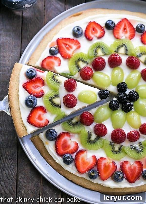 An overhead shot of a completed Fresh Fruit Pizza on a metal pan, with a single slice being removed, showcasing the perfect crust, frosting, and colorful fruit arrangement.