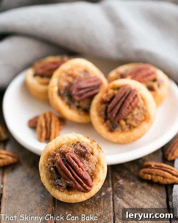 Pecan Tassies - Mini Pecan Pies in cream cheese pastry crusts. Rich and delicious!! Enjoy these perfect bite-sized desserts. Pecan Tassies on a white plate with pecan halves, showcasing the golden-brown crust and rich filling.