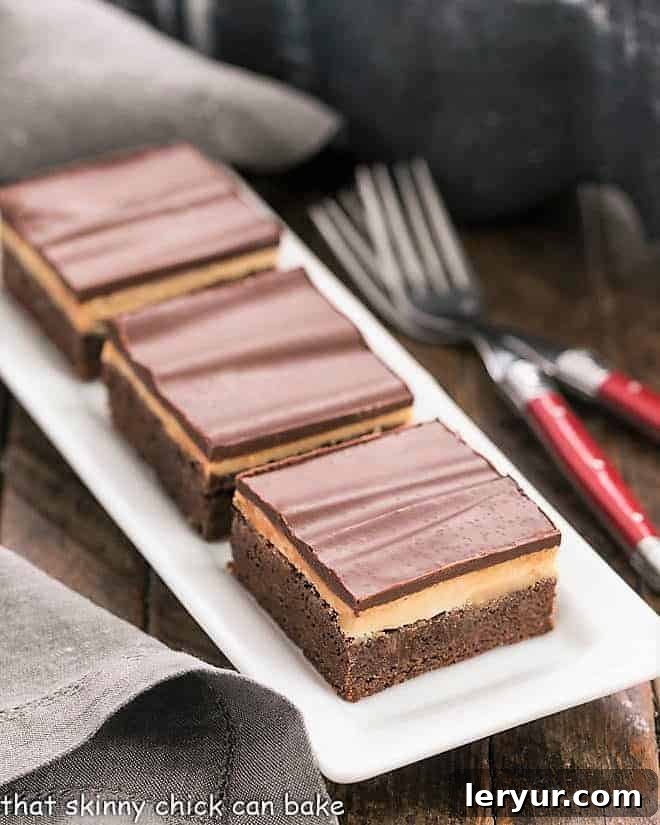 Peanut Butter Truffle Brownies - Fudgy brownies topped with a decadent peanut butter truffle filling and finished off with a dreamy chocolate ganache! Three decadent Peanut Butter Truffle Brownies stacked on a white ceramic tray, showcasing their distinct layers of fudgy chocolate, creamy peanut butter, and shiny ganache.