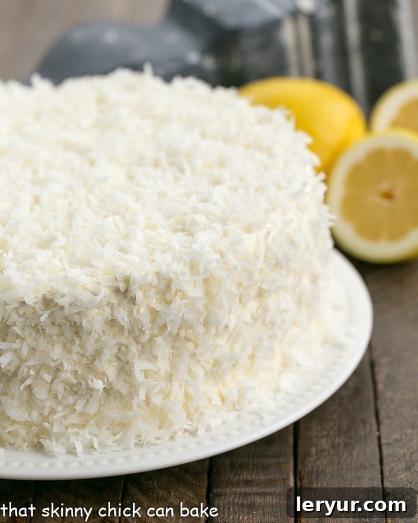 A stunning four-layer Lemon Layer Cake with Lemon Curd Filling, generously frosted with bright Lemon Swiss Meringue Buttercream and delicately topped with toasted coconut, presented on an elegant white serving plate, awaiting slicing.