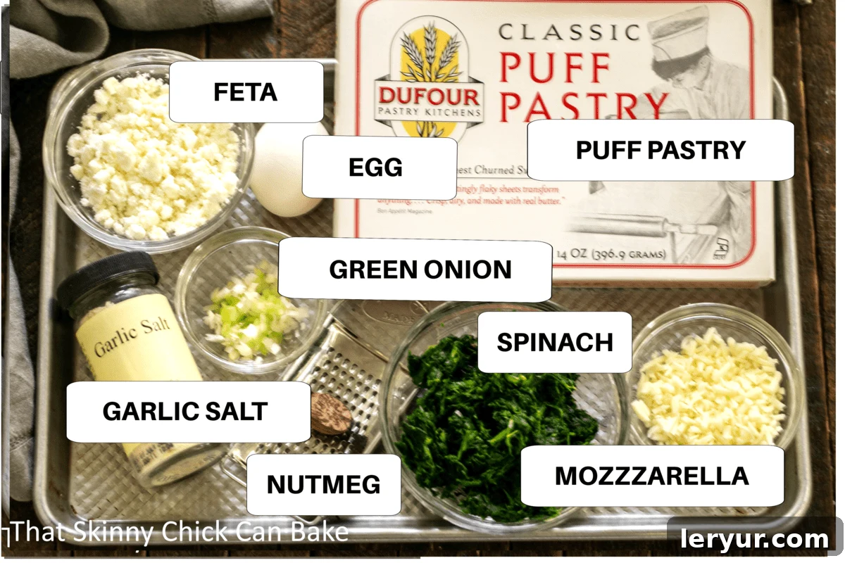 Golden Feta and Spinach Swirls 2 Carefully arranged ingredients for Spinach Feta Pinwheels on a metallic sheet pan, highlighting fresh spinach, feta block, mozzarella, green onions, and spices.