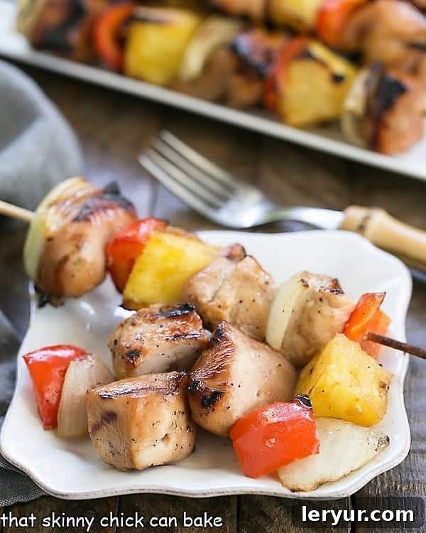 Honey Glazed Chicken Kabobs - Marinated chicken, veggies and pineapple are grilled to perfection! Plated Honey Glazed Chicken Kabobs on a small white plate, with a larger serving platter of kabobs in the background.