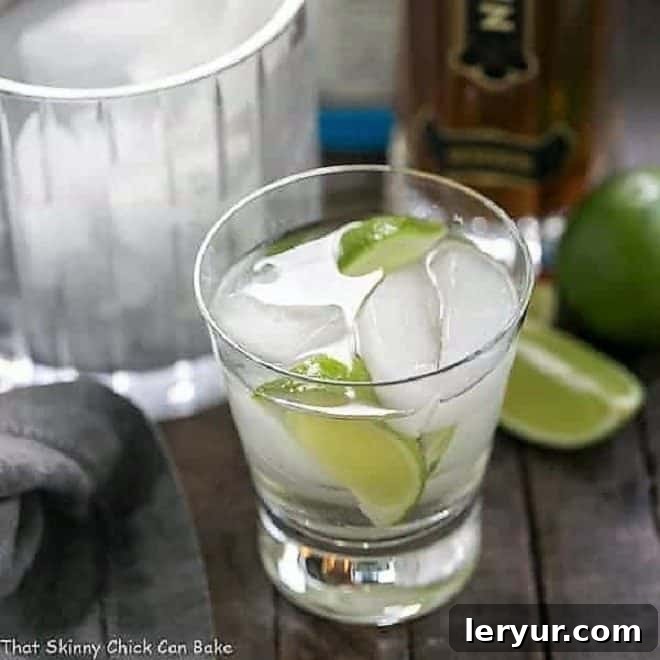 A single St. Germain Gin and Tonic Cocktail in a short, elegant glass, adorned with vibrant lime wedges and melting ice cubes, set against a blurred background.