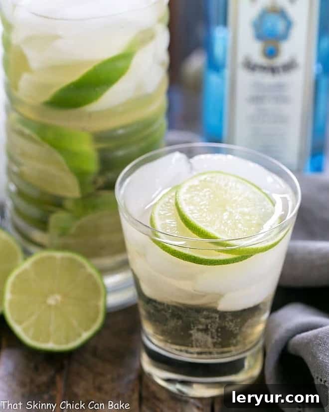 A beautifully presented St. Germain Gin and Tonic Cocktail in a short, clear glass, filled with ice cubes and adorned with multiple slices of fresh lime, on a light background.