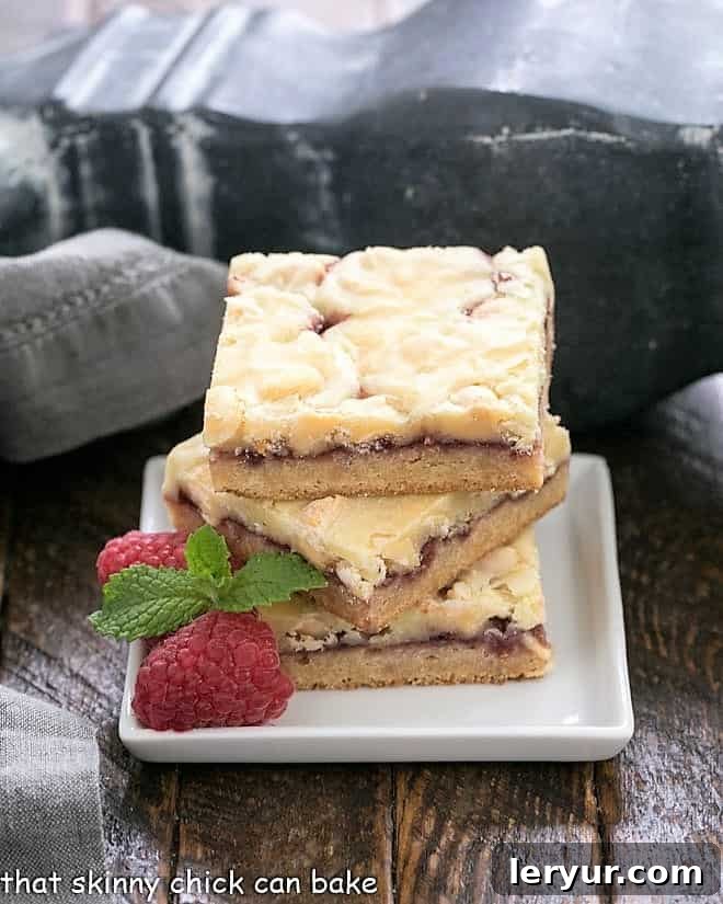 Decadent White Chocolate Raspberry Delight 3 A stack of white chocolate raspberry bars on a square white plate with raspberries and mint to garnish.