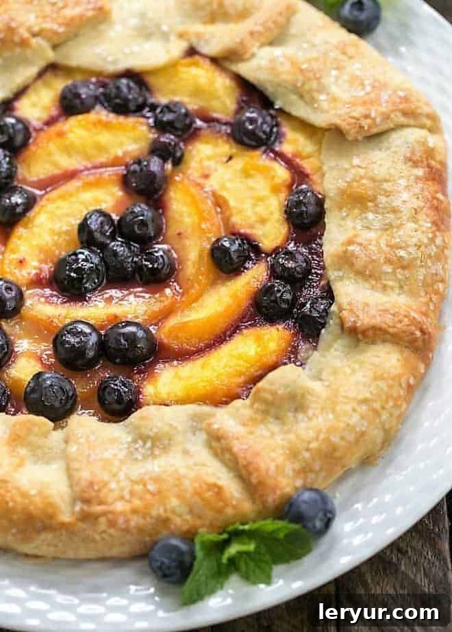 Peach Blueberry Galette - A gorgeous summer dessert with all the flavors of pie without all the work! A close-up view of a baked Peach Blueberry Galette, garnished with fresh blueberries and a sprig of mint, highlighting its flaky crust and juicy filling.