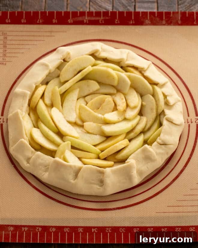 Unbaked apple galette on a sheetpan, with apples neatly arranged on the crust and edges folded over to contain the juices, ready for the oven.