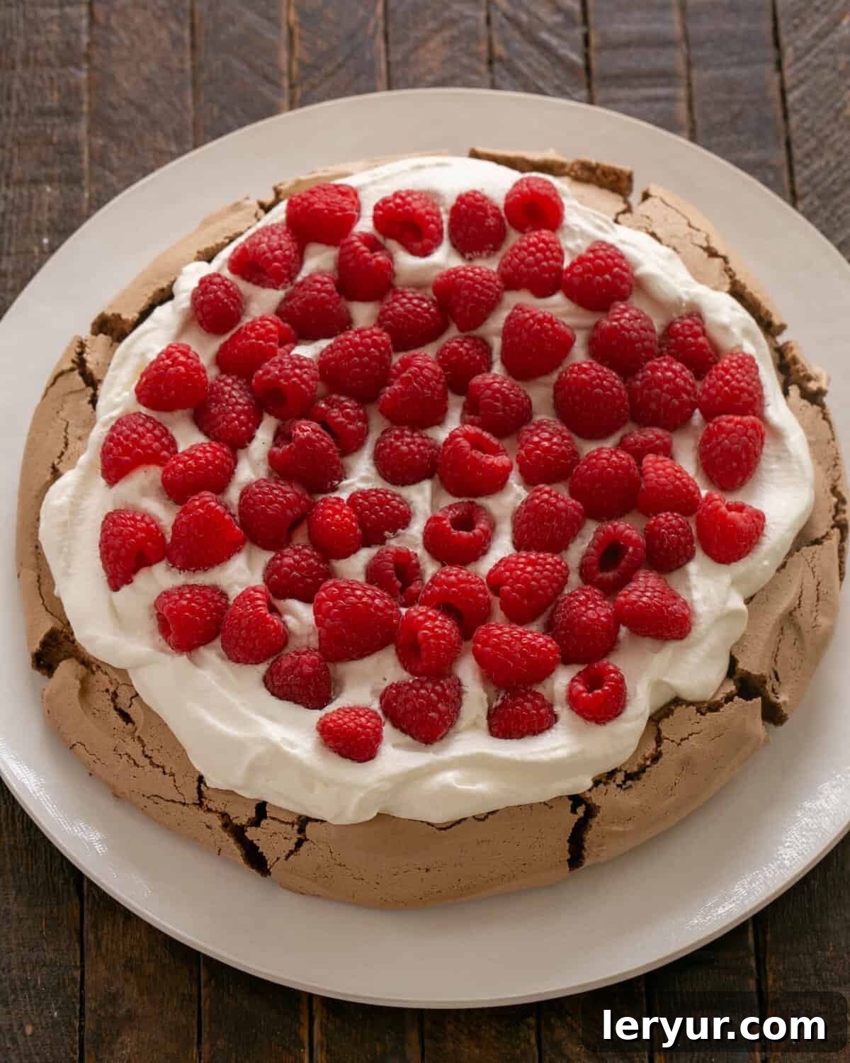 A cooled chocolate pavlova shell generously topped with sweetened whipped cream and fresh raspberries, ready for serving.
