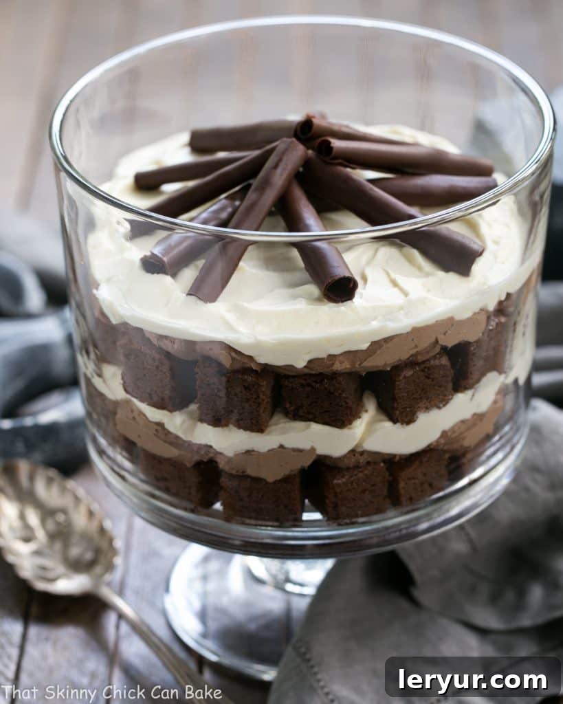 A beautifully presented Fudgy Brownie Trifle with Chocolate Mousse in a clear glass trifle dish, ready to be served with a silver spoon. The layers of dark brownie, light vanilla cream, and rich chocolate mousse are visible, topped with delicate chocolate curls.