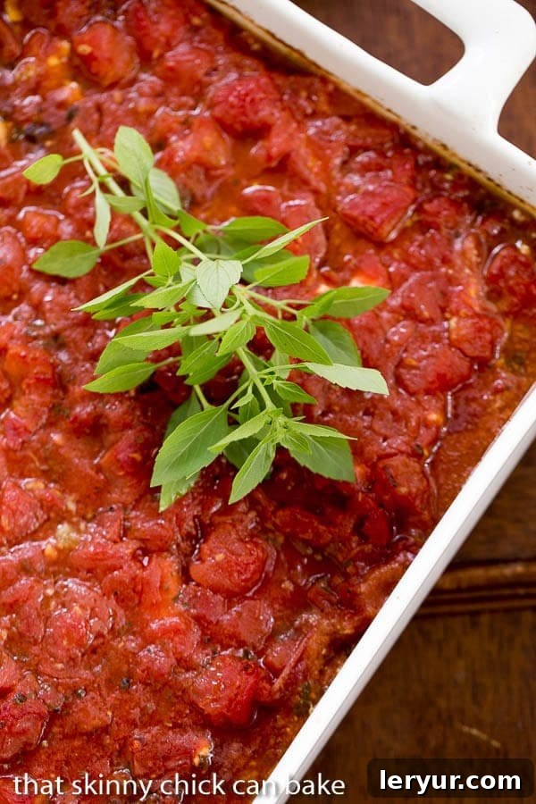 Chicken Feta Casserole - A fabulous casserole chock full of tomatoes, garlic, feta and herbs! A stunning close-up of Chicken Feta Casserole in a white casserole dish with a vibrant sprig of fresh basil, ready to be served.