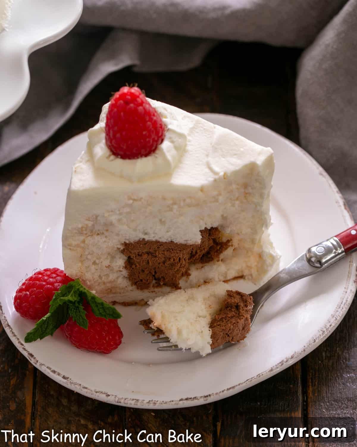 A stunning slice of mousse-filled angel food cake on a white plate, revealing a luscious chocolate center, garnished with fresh raspberries and a fork ready to enjoy.