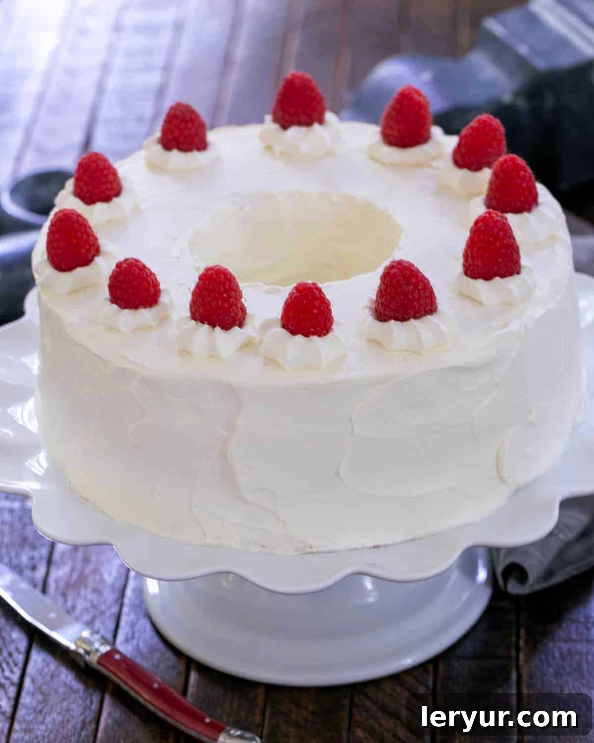 A stunning, perfectly frosted angel food cake with a visible chocolate mousse filling, beautifully garnished with fresh, ripe raspberries on a pristine white serving plate.