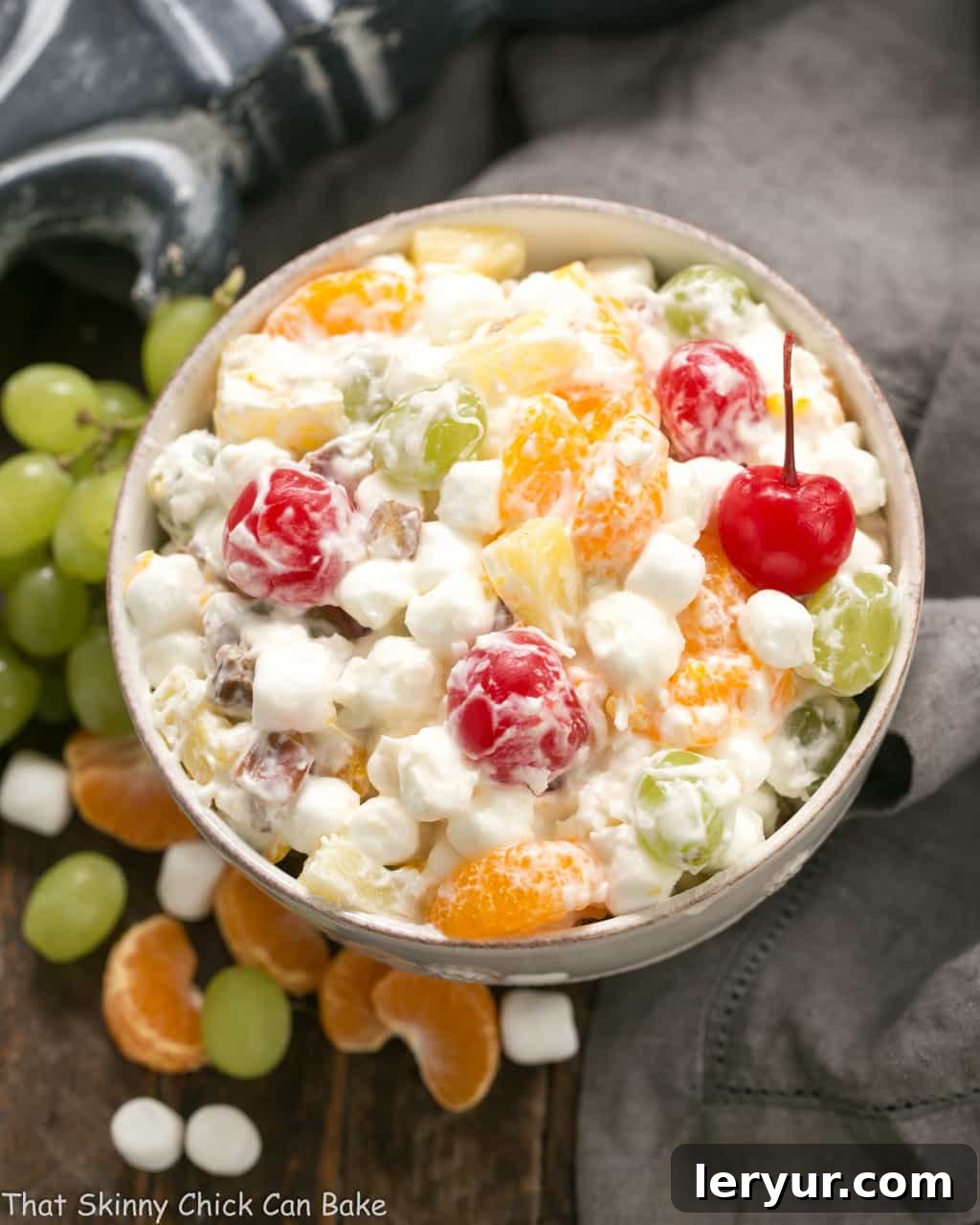 A large white ceramic bowl of Ambrosia Salad surrounded by its key ingredients: grapes, marshmallows, and vibrant orange segments.