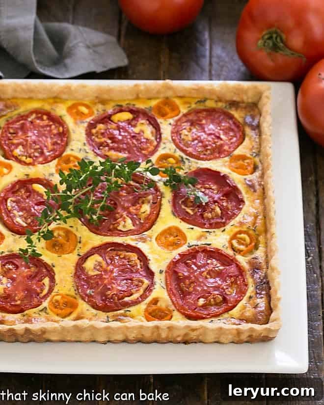 Tomato Gruyere Tart - A tasty way to enjoy the best of summer's tomatoes Overhead view of a beautifully golden-baked Tomato Gruyere Tart on a square white platter, garnished with fresh herbs, ready to be served.