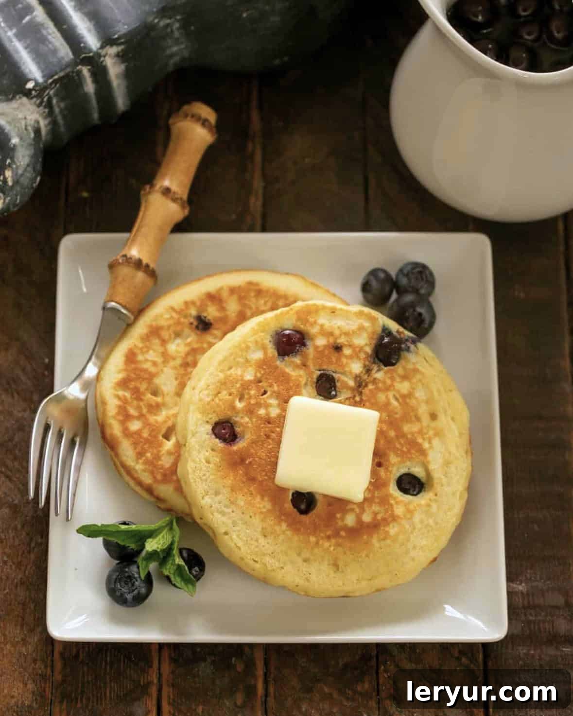 Buttermilk Pancakes with Blueberry Syrup - the quintessential pancake recipe with a blueberry infused maple syrup Two fluffy blueberry buttermilk pancakes on a square white plate with a bamboo handle fork, garnished with fresh blueberries.