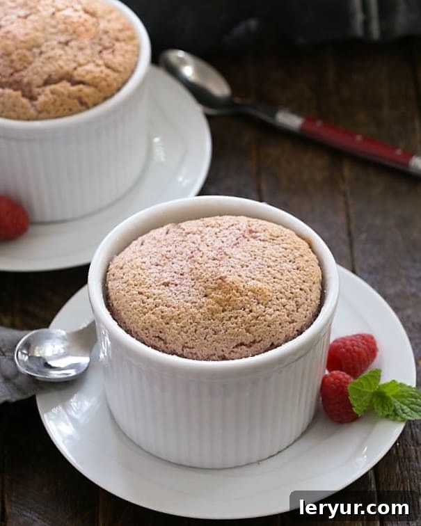 Two perfectly risen individual raspberry souffles, served on elegant white dishes with spoons, showcasing their delicate texture and vibrant color.