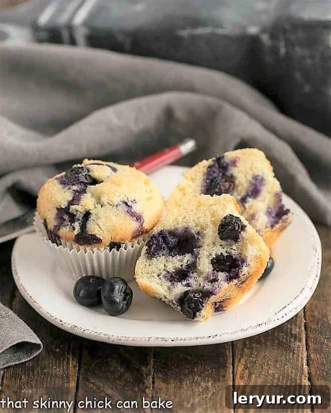 Blueberry Sour Cream Muffins - light, tender and chock full of juicy blueberries A beautifully presented plate of Sour Cream Blueberry Muffins, with one muffin elegantly cut in half, showcasing its moist, tender interior studded with juicy blueberries. This image emphasizes the inviting texture and deliciousness of the muffins.