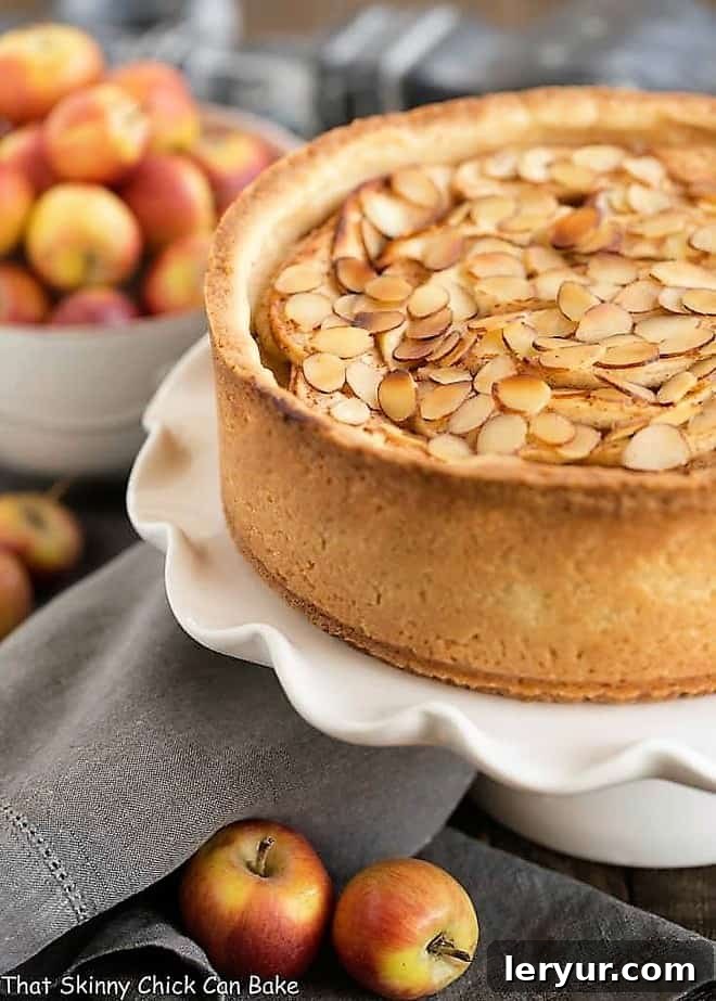 Cream Cheese Apple Torte - A buttery crust is topped with sweetened cream cheese, loads of cinnamon spiced apples, then sliced almonds A top-down view of the Cream Cheese Apple Torte on a ruffled white ceramic cake stand, garnished beautifully with lady apples, highlighting the toasted almonds and spiced apple layers.