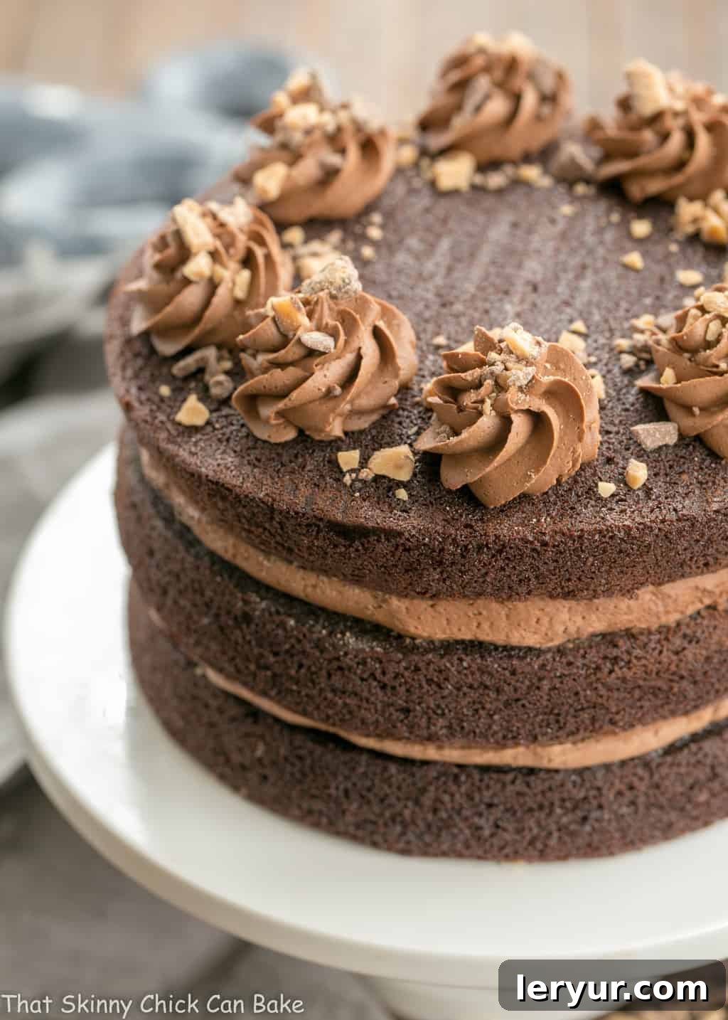 A stunning Triple Layer Chocolate Toffee Cake presented on a pristine white cake stand, showcasing its perfectly even layers of chocolate cake and rich buttercream frosting with toffee pieces visible.