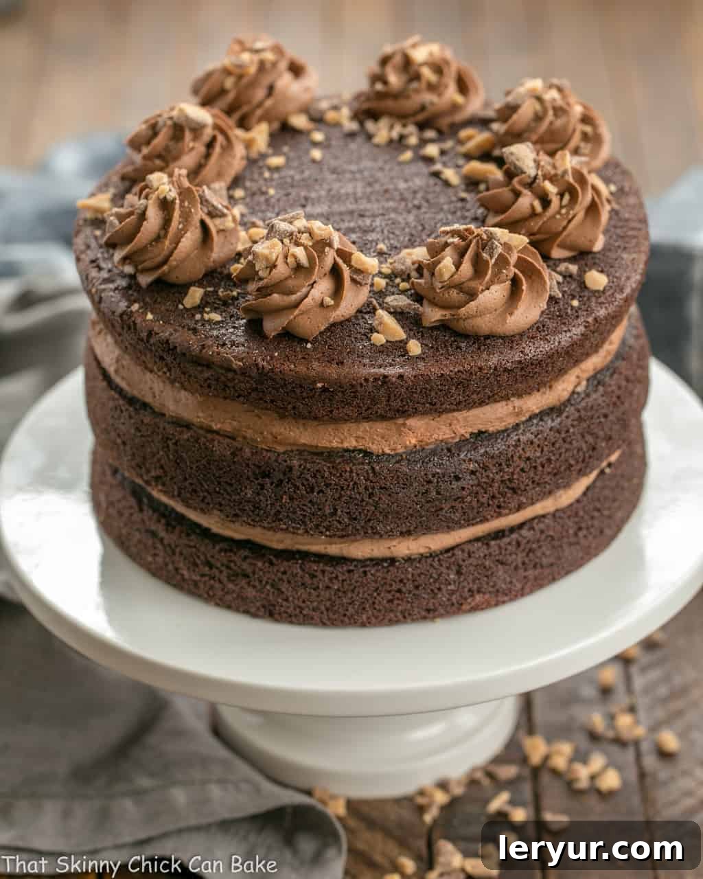 A full side view of the Triple Layer Chocolate Toffee Cake, showcasing its impressive height and layers, sitting on a white ceramic cake stand with a slice already removed.
