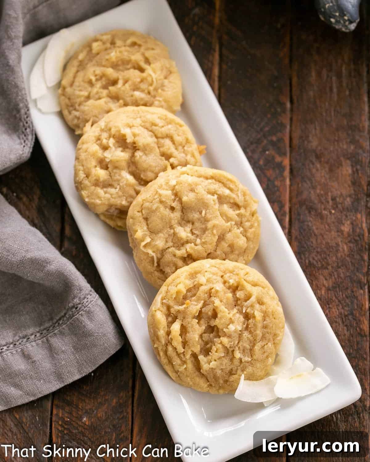 Perfectly Chewy Coconut Treats 2 Overhead view of 4 golden-brown chewy coconut cookies on a white ceramic tray, showcasing their perfect texture and toasted coconut flecks.