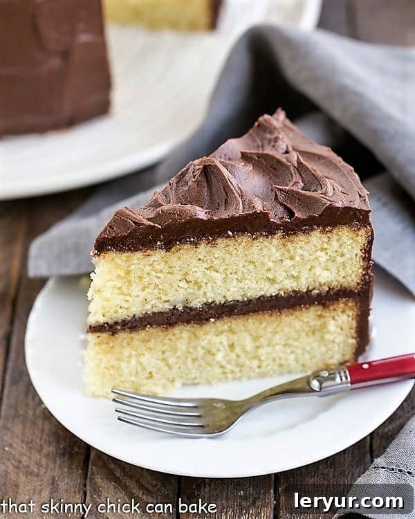 Perfect Yellow Cake Recipe with Chocolate Buttercream - An exquisite, tender yellow cake topped with dreamy chocolate frosting! A delectable slice of Perfect Yellow Cake Recipe with Chocolate Buttercream, served on a white plate with a charming red-handled fork, inviting indulgence.