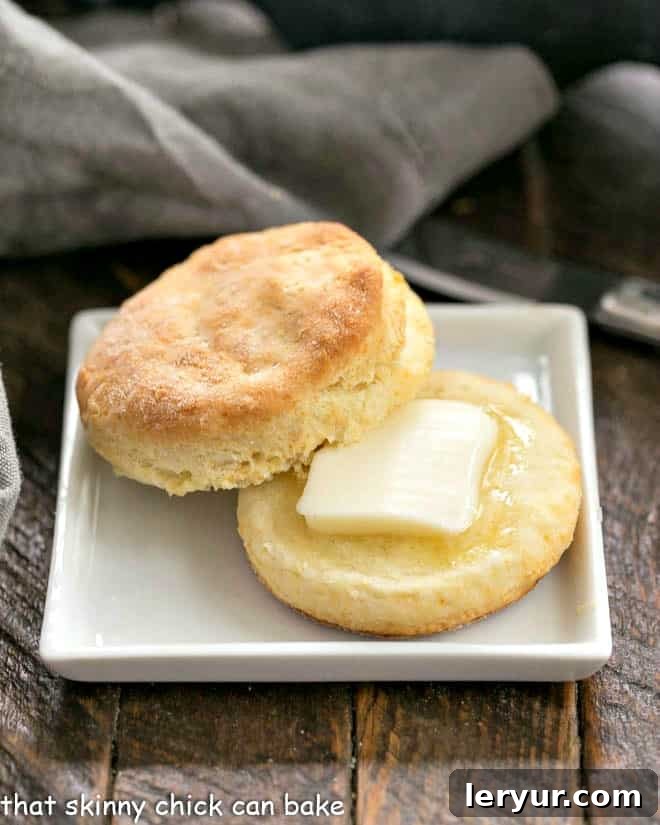 A perfectly split homemade buttermilk biscuit, revealing its intricate flaky layers, with a pat of butter gently melting into its warm, inviting interior. This culinary delight is presented on a square white plate, ready to be savored.